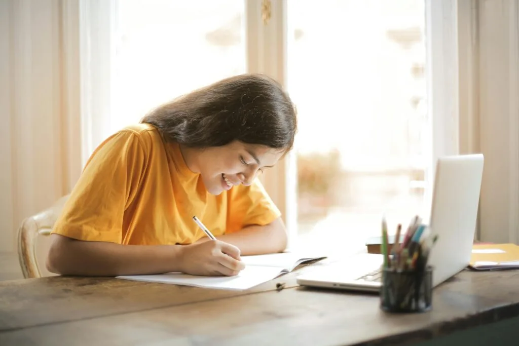 pexels-photo-3807755-3807755 Young woman studying with a laptop and notepad at home, smiling and focused.