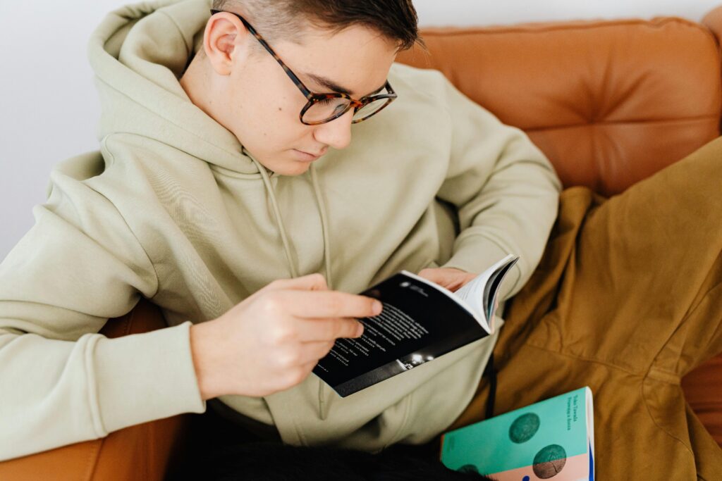 pexels-photo-6958699-6958699 A teenager in glasses and a hoodie reading a book on a comfortable couch indoors.