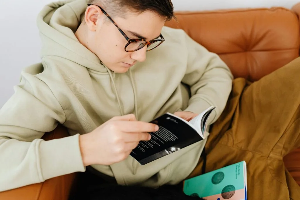 A teenager in glasses and a hoodie reading a book on a comfortable couch indoors.